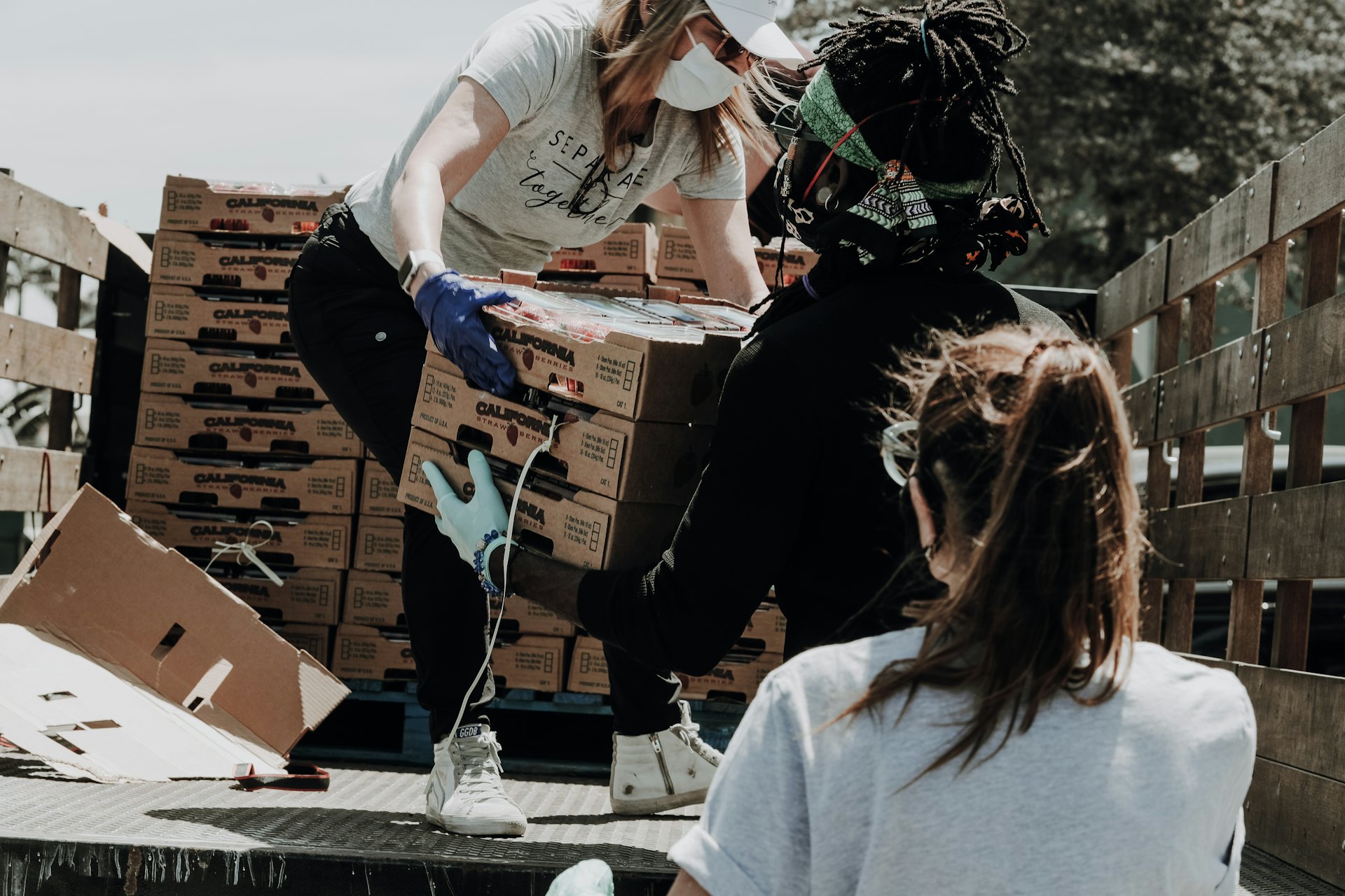 Three people wearing masks loading cardboard boxes of strawberries onto a truck bed